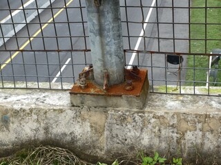 Close-up detail of a structural post base plate and anchor bolts heavily affected by rust, secured to a concrete barrier on an overpass above a highway.