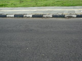Low-angle view of an asphalt road meeting a sidewalk with black and white painted curb blocks, bordered by a green grass verge above.