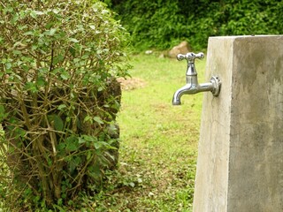 A bright chrome faucet installed on a rough concrete pillar in a lush green outdoor setting, with a dense green bush on the left.