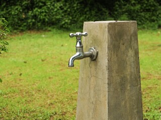 A bright chrome garden faucet installed on a tall concrete pillar. The fixture stands in a lush green grassy area with dense foliage in the background.