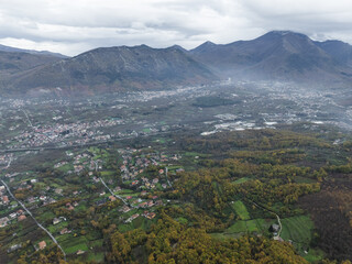 Aerial view of a quaint town nestled amidst rolling hills, with a muted palette under a cloudy sky, the landscape a blend of autumnal hues and verdant evergreens, Serino, Irpinia, Italy.