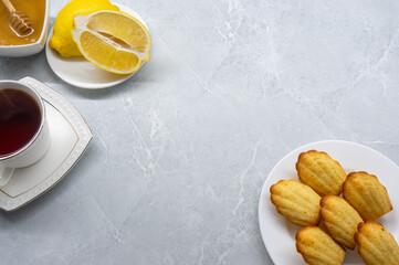Madeleine cookies on a white plate with lemon, honey, and a cup of tea against a light background. Space for text.