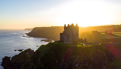 Obraz premium Aerial view captures a sunlit, medieval fortress perched atop a rugged cliff overlooking a vast ocean. Golden hour bathes the scene