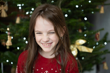 Girl in red Christmas sweater with white snowflakes smiling and looking down, visible teeth near decorated tree indoors