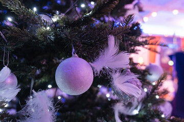 Close up of white christmas ball ornament with feathers and lights on decorated tree in elegant festive interior with blue bokeh background