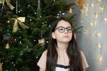 Sad teenage girl in glasses and T-shirt near decorated Christmas tree looking upward sideways, emotional holiday childhood concept