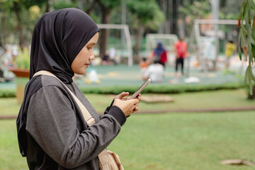 A young Indonesian Muslim woman typing a message on her smartphone in a public park