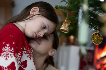 Portrait of mother hugging sad daughter in Christmas sweater near decorated tree, emotional holiday family moment