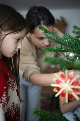 Girl in red Christmas sweater standing near mother decorating tree with handmade ornaments in foreground, cozy family holiday moment