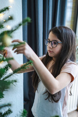 Teenage girl in glasses and T-shirt decorating Christmas tree at home, cozy holiday family moment