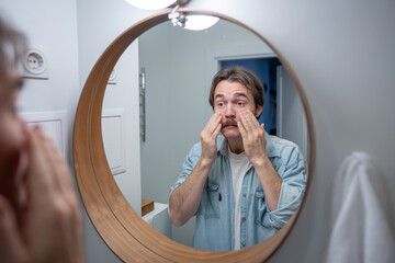 Thoughtful unhappy man looking at reflection in bathroom mirror touching tired face. Skin fatigue, signs of stress, lack of sleep. Dissatisfaction with appearance, low self esteem, body image issues.