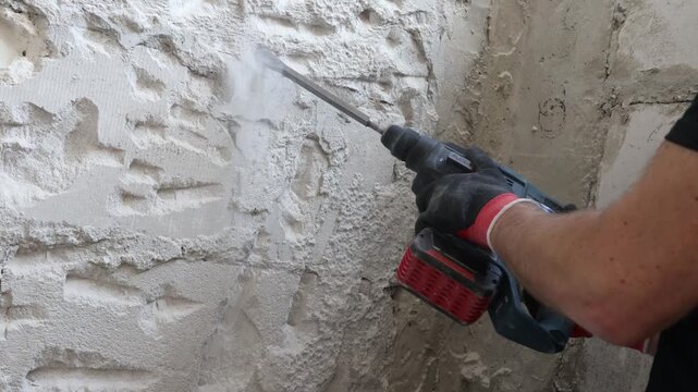 Close-up of worker using electric hammer drill to remove old plaster from concrete interior wall. Renovation and demolition process with rough wall texture, dust and damaged surface. Concept of home i