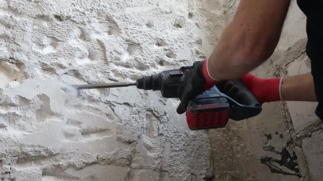 Close-up of worker hands using electric hammer drill to remove old plaster from interior concrete wall. Renovation and demolition process with rough texture, dust and damaged surface. Concept of home 