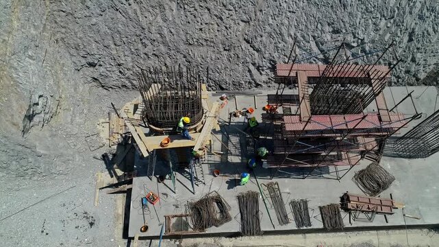 Top-down aerial view of workers installing rebar and formwork for a reinforced concrete bridge pier foundation. Civil engineering work at a large highway infrastructure construction site.