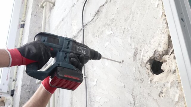 Close-up of construction worker drilling concrete wall using rotary hammer drill. Electrical installation, cable routing, renovation and repair work at construction site. Professional building and ind