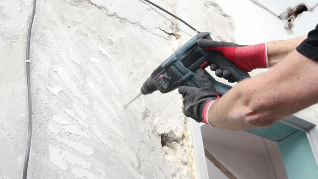 Close-up of construction worker drilling concrete wall using rotary hammer drill. Electrical installation, cable routing, renovation and repair work at construction site. Professional building and ind