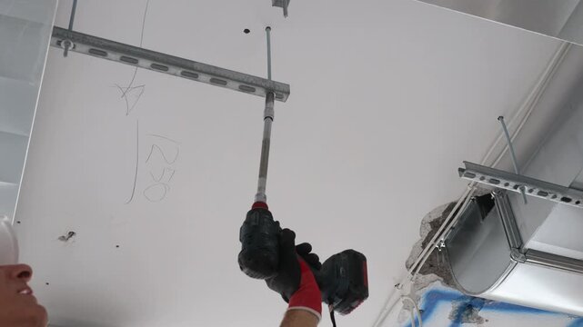 Construction worker installing metal frame for drywall ceiling using cordless drill. Professional interior renovation process, ceiling mounting system, metal profiles installation, home improvement, b