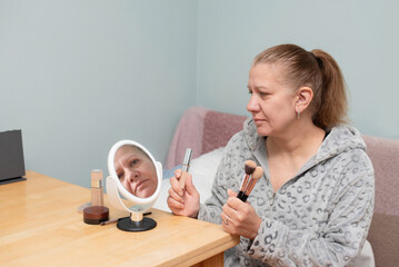 A middle-aged woman is about to do her morning make up, has prepared brushes, sponges, foundation and lipstick, looks at herself in the mirror and regrets her young years