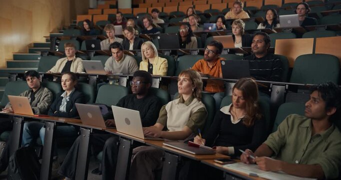 Diverse Group of University Students of Various Ethnicities Sit in Lecture Theater, Listening to an Unseen Professor While Typing on Laptops, Higher Education Classroom Presentation.
