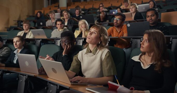 Diverse Group of University Students Sit in a Large Lecture Hall, Typing on Laptops, Taking Handwritten Notes, and Concentrating on an In-depth Academic Presentation and Higher Education Coursework.