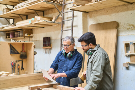 Experienced craftsman teaching young apprentice in busy lumberyard workshop