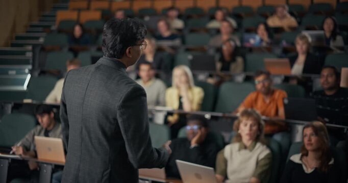 Professor in a Gray Suit Delivers an Interactive University Lecture, Gesturing Toward a Diverse College Audience Using Laptops, Engage in Higher Education Learning.