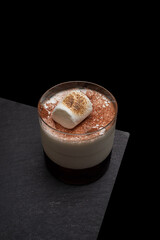 A delicious cocktail garnished with a toasted marshmallow and cocoa powder, artfully presented on a black surface. The drink looks appetizing and elegant.