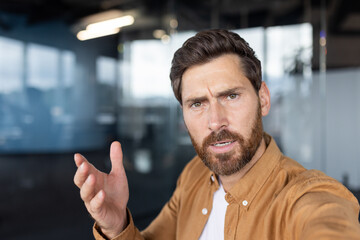 Fototapeta premium Confused man experiencing frustration and asking questions while shrugging and talking during a video call or online meeting in a modern corporate office setting