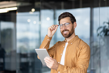 Confident young businessman in a modern office holding a tablet, adjusting his glasses and smiling...