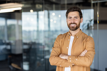 Smiling bearded man standing in a modern office with arms crossed, projecting confidence,...