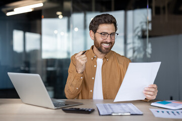 Happy businessman looking at a document and celebrating an achievement, showing success and good news in his office workspace with a laptop and papers on the desk