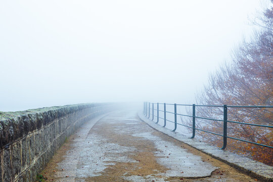 Camino entre la niebla con muro de piedra y barandilla met&aacute;lica