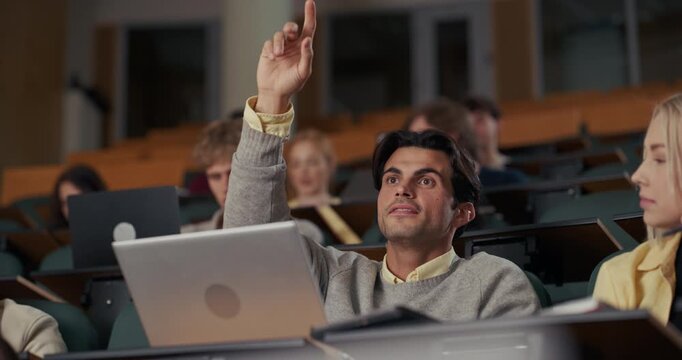 Male Student in a Crowded University Lecture Hall Types on Laptop, Then Confidently Raises His Hand to Answer the Professor&rsquo;s Question, Interactive Learning While Diverse Classmates Listen