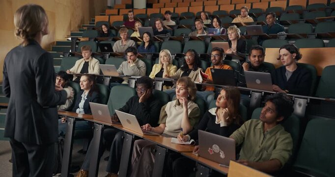Female Professor Leads an Interactive University Lecture as a Diverse Group Students With Laptops Listen, Take Notes, Raise Hands, Showcasing Active Higher Education Learning, Classroom Engagement.
