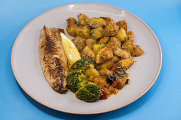 Grilled mackerel fillet served with spiced roasted potato wedges and sautéed vegetables, presented on a plain blue background. Healthy homemade Mediterranean-style meal
