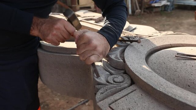Close up of artisan hands carving stone with chisel and hammer. Skilled sculptor creating detailed architectural ornament. Traditional manual craftsmanship process in a workshop setting.

