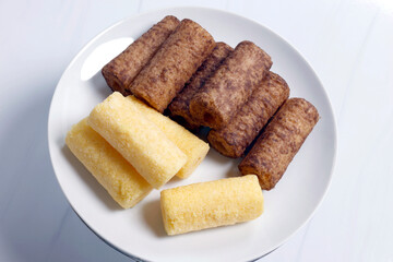 Assorted baked snacks on a white plate, including chocolate sticks, milk flavored sticks, and a small cake. Ideal for food, bakery, and dessert concepts, Top view