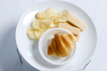 White plate with assorted snacks including a croissant, potato chips, and wafer sticks. Clean and simple setup suitable for food menus, Top view