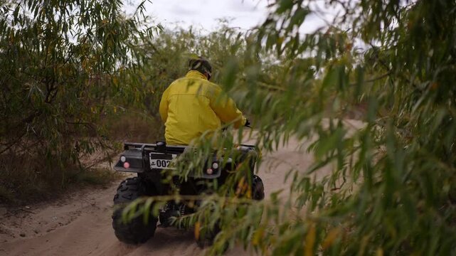 man in yellow jacket riding ATV down sandy trail, dust kicking up behind tires, dense green shrubs lining narrow path, serene overcast sky, engine rumble