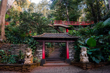 Torii style gate with stone guardian lions leads up steps under a red lattice footbridge in Monte Palace Tropical Garden, Funchal, Madeira, Portugal, in soft daylight.