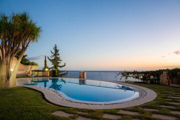 Curved infinity pool on a terrace in Madeira near Funchal faces the Atlantic at dusk. Cypress and dragon trees reflect, garden lights glow, stone and pebbles line the edge.