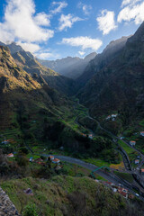 Sunlight filters onto a deep green valley in Madeira, Portugal, with terraced fields, white roofs, a winding road, a small bridge, and a river leading toward misty peaks.