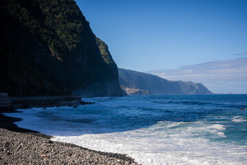 Steep forested cliffs rise over a pebble beach and Atlantic surf on Madeira's north coast, Portugal. Terraced villages dot slopes, a headland curves, midday light defines