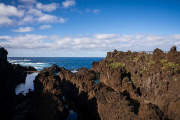 Jagged lava rocks surround calm tidal pools at Piscinas Naturais do Porto Moniz, Madeira. Waves crash beyond the basalt barrier under crisp daylight and scattered clouds.