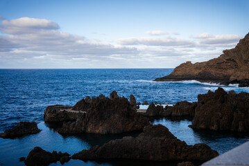 Jagged lava rocks form seawater pools at Porto Moniz, Madeira, Portugal, as Atlantic waves break against a headland under soft clouds in late afternoon cool light.