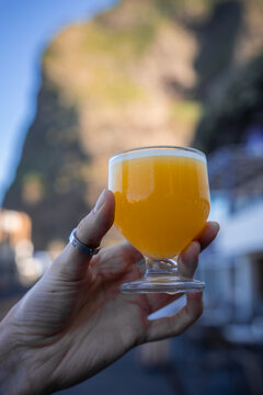 A hand holds a stemmed glass of bright orange frothy drink. Warm afternoon light and shallow depth of field frame sunlit cliffs and coastal buildings in Madeira, Portugal.