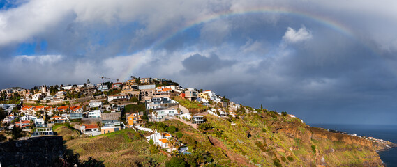 A rainbow arcs above terraced villas on a rugged coastal cliff in Funchal, Madeira Island, Portugal. Golden light meets dark clouds as the Atlantic and basalt cliffs frame the view.