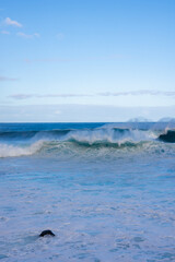 Powerful Atlantic waves roll toward shore at Madeira, Portugal, windblown spray rising as foamy water churns, a lone dark rock visible, soft daylight and minimalist frame.