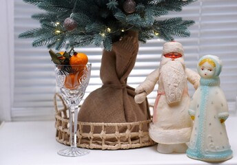 vintage figures of Soviet Santa Claus and Snow Maiden near a retro crystal glass with tangerines under a Christmas tree on a white windowsill