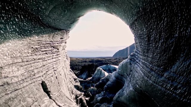 Cinematic aerial drone flight through Katla ice cave tunnel showing blue ice with black volcanic ash veins and green mossy mountains in Iceland 4K.